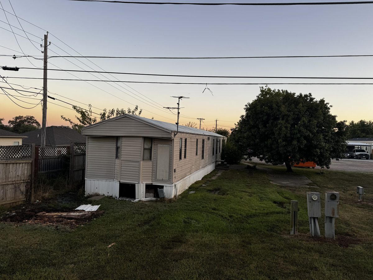 Mobile home waiting on utility disconnect before demolition in Columbia, MO, with the overhead electric drop still attached and meter pedestals standing in the yard